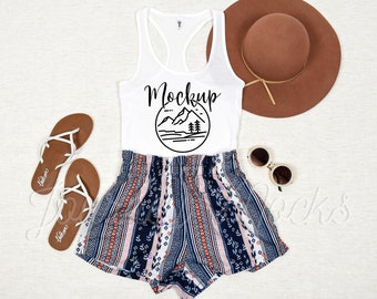 May include: White tank top with a black mountain graphic and the word "Mockup" printed on the front. The tank top is paired with a pair of blue and white striped shorts and a brown hat.