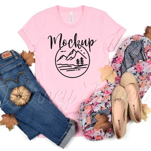 May include: Pink t-shirt with a black mountain graphic and the word "Mockup" printed on the front. The shirt is lying on a white surface with a pair of jeans, a floral scarf, and autumn leaves.