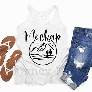 May include: White tank top with a black graphic of mountains and trees in a circle with the word "Mockup" above it. The tank top is laying flat on a white background with a pair of brown sandals and a pair of blue jeans.