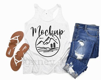 May include: White tank top with a black graphic of mountains and trees in a circle with the word "Mockup" above it. The tank top is laying flat on a white background with a pair of brown sandals and a pair of blue jeans.