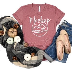 Op de afbeelding: Een mauve V-hals T-shirt met een witte afbeelding van bergen en bomen in een cirkel met het woord "Mockup" erboven. Het shirt ligt op een wit oppervlak met een geruite deken, een blauwe jeans en een paar beige ballerina's.