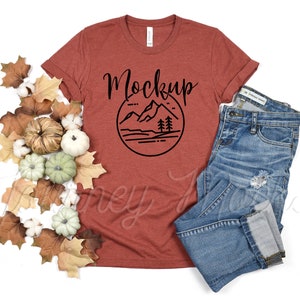 May include: A rust-colored t-shirt with a black mountain graphic and the word "Mockup" in a handwritten font. The t-shirt is laying on a white surface with fall leaves and cotton balls around it. A pair of blue jeans is also laying on the surface.