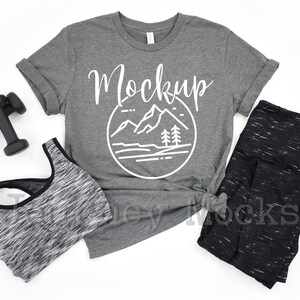 May include: A heather gray t-shirt with a white mountain graphic and the word "Mockup" printed on the front. The shirt is laid flat on a white surface with a black and gray sports bra, black and gray leggings, and a pair of black dumbbells.