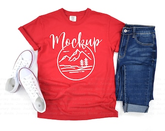 May include: A red t-shirt with the word "Mockup" and a mountain and tree graphic in white. The shirt is paired with blue jeans and white sneakers, creating a casual outfit.