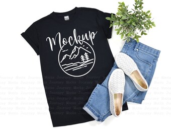 Puede incluir: Una camiseta negra con un gráfico de montaña blanco y la palabra "Mockup" impresa en ella. La camiseta está colocada sobre una superficie blanca con un par de pantalones vaqueros azules y zapatos blancos.