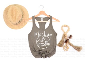 May include: A mockup of a sage green tank top with a white graphic of a mountain range and trees inside a circle. The text "Mockup" is above the graphic. The tank top is hanging on a wooden hanger. There is a straw hat, wooden beads, and sunglasses in the image.
