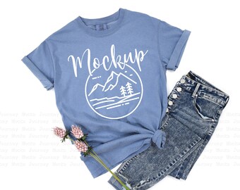 May include: A light blue t-shirt with a white graphic of mountains and trees in a circle. The text "Mockup" is written in white above the graphic.