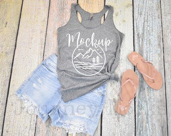 May include: A heather grey tank top with a white mountain graphic and the word "Mockup" printed on the front. The tank top is laying on a wooden surface with a pair of denim shorts and brown flip flops.