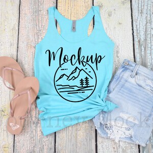 May include: A turquoise tank top with a black graphic of mountains and trees in a circle. The text "Mockup" is written in black cursive above the graphic. The tank top is laying on a light wood surface with a pair of brown flip flops and a pair of light blue denim shorts with lace trim.