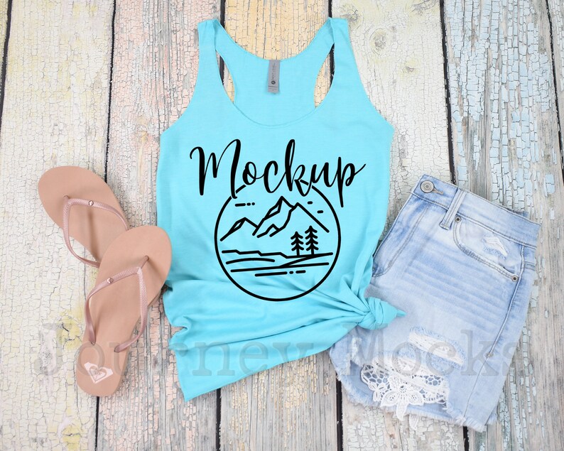 May include: A turquoise tank top with a black graphic of mountains and trees in a circle. The text "Mockup" is written in black cursive above the graphic. The tank top is laying on a light wood surface with a pair of brown flip flops and a pair of light blue denim shorts with lace trim.