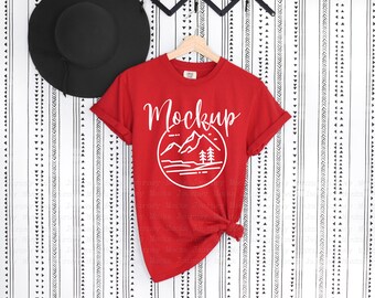 May include: Red t-shirt mockup with a white graphic of mountains and trees in a circle with the word "Mockup" above it. The shirt is hanging on a hanger against a white and black patterned background.