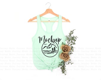 Op de afbeelding: Een mintgroen tanktop met een zwarte afbeelding van een bergketen en het woord "Mockup" op de voorkant. Het tanktop hangt aan een houten hanger.
