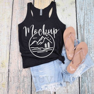 May include: Black tank top with a white graphic of mountains and trees. The text "Mockup" is written in white on the front of the tank top. The tank top is laying on a wooden surface with a pair of brown flip flops and denim shorts.