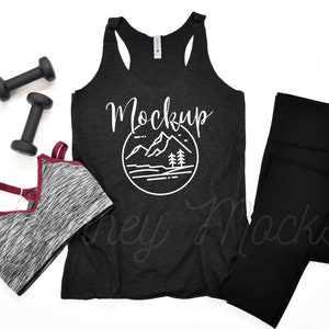 May include: Black racerback tank top with a white mountain graphic and the word "Mockup" in white text. The tank top is laying flat on a white surface with a black sports bra, black leggings, and two black dumbbells.