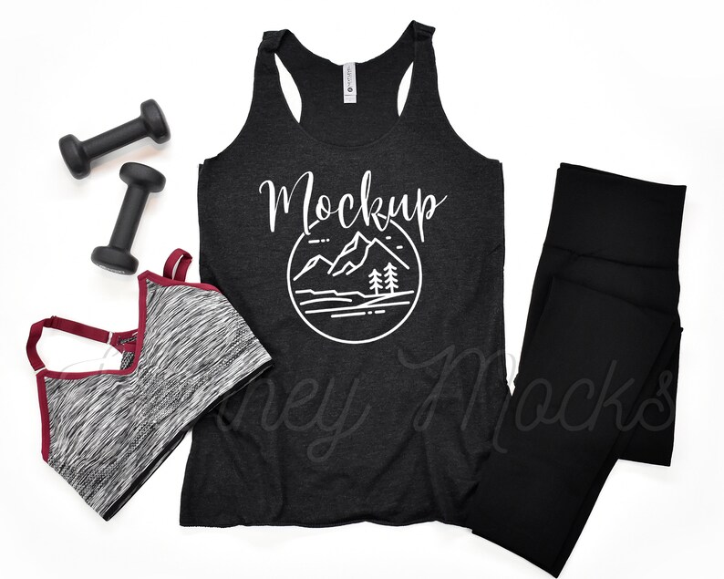 May include: Black racerback tank top with a white mountain graphic and the word "Mockup" in white text. The tank top is laying flat on a white surface with a black sports bra, black leggings, and two black dumbbells.