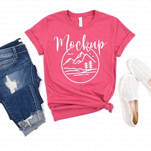 May include: A pink t-shirt with a white graphic of mountains and trees in a circle. The text "Mockup" is above the graphic. The t-shirt is laid flat on a white background with a pair of blue jeans and white striped slip-on shoes.