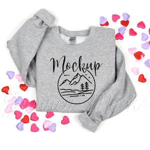 Puede incluir: Sudadera gris con una escena de montaña y la palabra "Mockup" impresa en la parte delantera. La sudadera está rodeada de corazones rosas y morados.