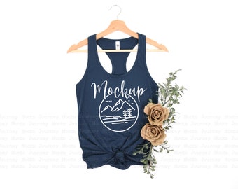 Op de afbeelding: Een marineblauw tanktop met een witte afbeelding van een bergketen en het woord "Mockup". Het tanktop hangt aan een houten hanger en is gestyled met een takje groen en twee beige rozen.