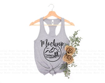 May include: A gray tank top with a black mountain graphic and the word "Mockup" printed on the front. The tank top is hanging on a wooden hanger and is styled with a sprig of greenery and two beige flowers.