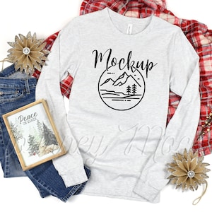 Op de afbeelding: Een wit longsleeve t-shirt met een zwarte bergketen graphic en het woord "Mockup" in een handschrift. Het shirt ligt plat op een witte achtergrond met een rood-zwart geruit deken en een bruin stervormig ornament.