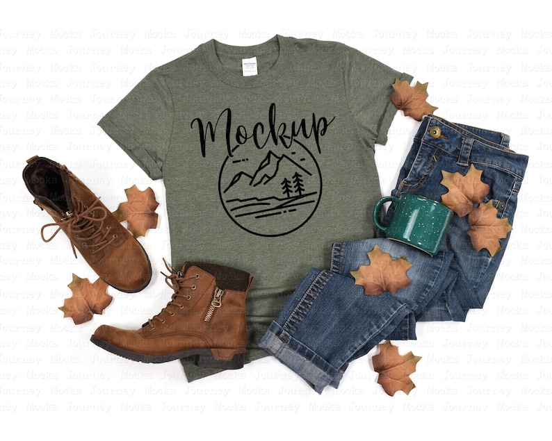May include: A green t-shirt with a black mountain scene graphic and the word "Mockup" in a handwritten font. The shirt is laid out on a white background with a pair of blue jeans, brown boots, and fall leaves.