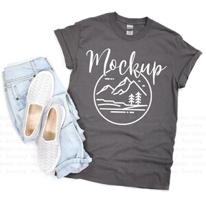 May include: A charcoal gray t-shirt with a white graphic of a mountain range and the word "Mockup" printed on the front. The t-shirt is laid flat on a white surface with a pair of white canvas shoes and blue jeans.