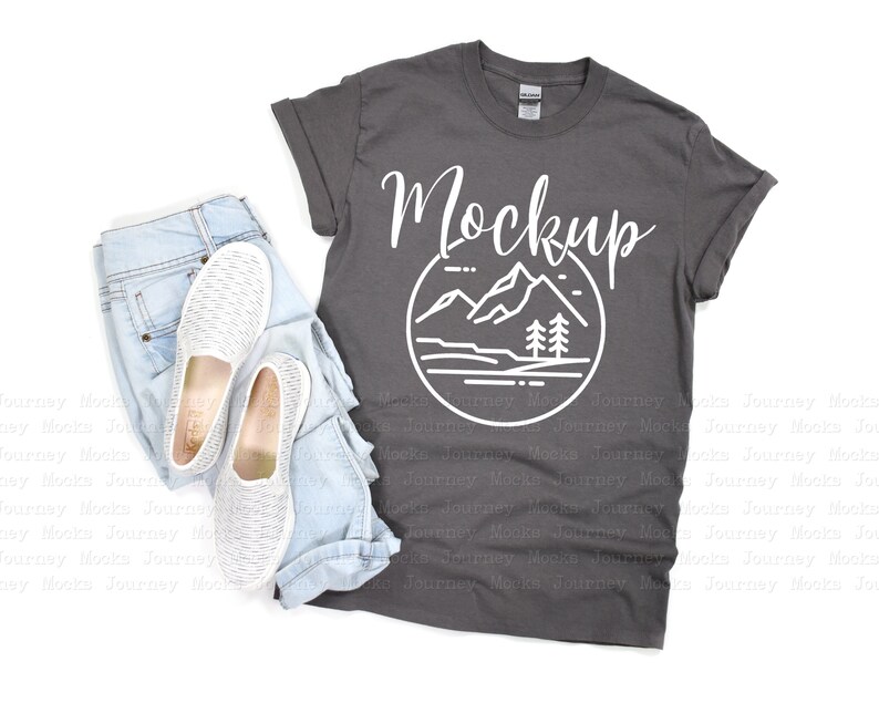 May include: A charcoal gray t-shirt with a white graphic of a mountain range and the word "Mockup" printed on the front. The t-shirt is laid flat on a white surface with a pair of white canvas shoes and blue jeans.