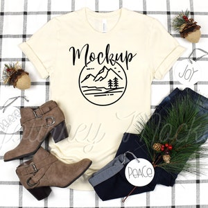 Op de afbeelding: Een beige T-shirt met een zwarte bergprint en het woord "Mockup" erop gedrukt. Het shirt ligt plat op een geruit oppervlak met een paar bruine enkellaarsjes, een blauwe jeans en kerstversiering.