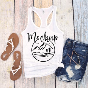 Könnte beinhalten: Ein weißes Tanktop mit dem Wort "Mockup" und einer Berg- und Baumgrafik. Das Tanktop wird mit Jeansshorts und braunen Sandalen mit weißen Riemen kombiniert. Die Artikel sind auf einer hellen Holzmaserung angeordnet.