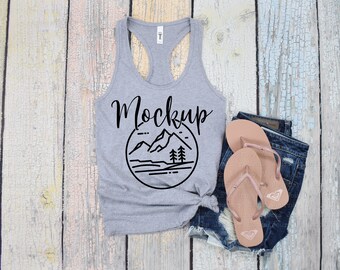 May include: Gray tank top with a black mountain and trees graphic and the word "Mockup" printed on the front. The tank top is laying on a light wood surface with a pair of brown flip flops and denim shorts.