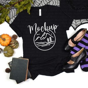 Puede incluir: Camiseta negra con un gr&aacute;fico de monta&ntilde;a blanco y la palabra "Mockup" en una fuente manuscrita. La camiseta est&aacute; sobre una superficie blanca con un sombrero de bruja, medias a rayas negras y moradas y zapatos negros.