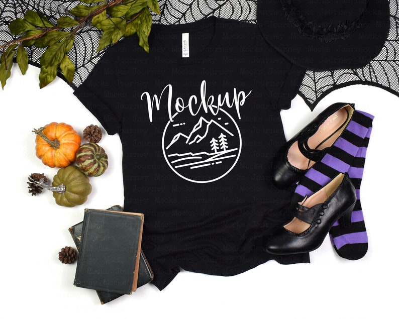 Puede incluir: Camiseta negra con un gr&aacute;fico de monta&ntilde;a blanco y la palabra "Mockup" en una fuente manuscrita. La camiseta est&aacute; sobre una superficie blanca con un sombrero de bruja, medias a rayas negras y moradas y zapatos negros.