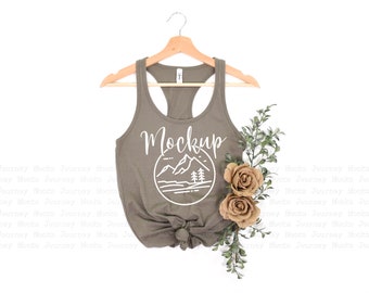 May include: A mockup of a sage green tank top with a white graphic of a mountain range and the word "Mockup" on the front. The tank top is hanging on a wooden hanger and is styled with a sprig of greenery and two beige roses.