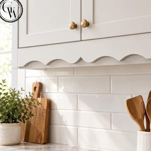 May include: Off-white kitchen cabinets with a scalloped bottom edge and round brass knobs. Below the cabinets is a white subway tile backsplash. A wooden cutting board, a small potted green plant, and wooden spoons are visible in the foreground.