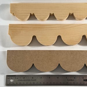 May include: Three wooden decorative trim pieces with a scalloped edge. The top two pieces are light-colored wood, and the bottom piece is a medium-brown composite material. A ruler is at the bottom of the image.