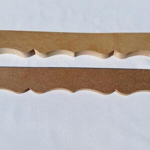 May include: Two unfinished wooden decorative trim pieces. The pieces are rectangular with a scalloped edge along the bottom. One piece is lighter in colour than the other. The pieces are laying on a white surface.