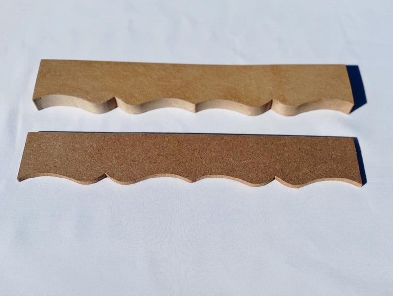 May include: Two unfinished wooden decorative trim pieces. The pieces are rectangular with a scalloped edge along the bottom. One piece is lighter in colour than the other. The pieces are laying on a white surface.