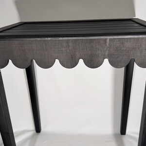 May include: A black wooden side table with a scalloped apron and a slatted top. The table has four straight legs and is painted in a dark, matte finish. The design is simple and elegant.