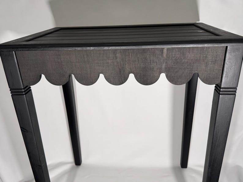 May include: A black wooden side table with a scalloped apron and a slatted top. The table has four straight legs and is painted in a dark, matte finish. The design is simple and elegant.