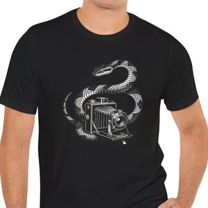 May include: A black short-sleeved t-shirt featuring a white graphic illustration of a vintage bellows camera coiled by a snake. The design is centered on the chest, showcasing the detailed lines of the camera lens and the snake's scales.