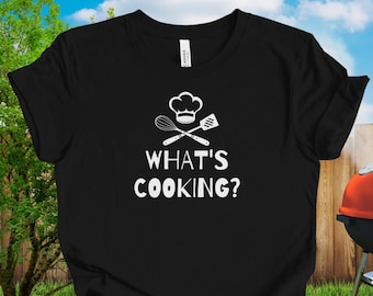 May include: Black t-shirt with the text "WHAT'S COOKING?" in white, along with a chef's hat, whisk, and spatula graphic. A red and black grill is in the background, set on a green lawn with a wooden fence and a tree.