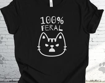 Puede incluir: Camiseta negra con el texto "100% FERAL" sobre una cara de gato blanco de dibujos animados. El gato tiene una expresión enfadada. Es una prenda informal.