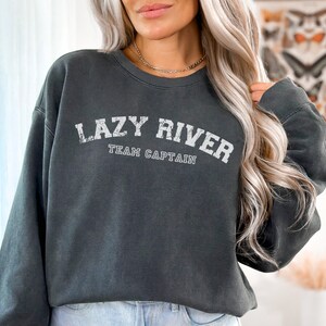 Puede incluir: Una sudadera gris oscuro con el texto "LAZY RIVER TEAM CAPTAIN" en letras blancas. La sudadera tiene un corte holgado y cuello redondo.