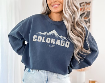Peut inclure: Un sweat-shirt bleu marine avec un motif blanc de montagnes et le texte "COLORADO Est. 1876".