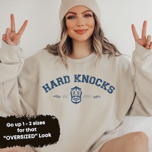 Könnte beinhalten: Ein beiger Pullover mit dem Text "HARD KNOCKS" in Blau mit einer blauen Grafik eines Löwenkopfes und dem Text "Est. 1984".
