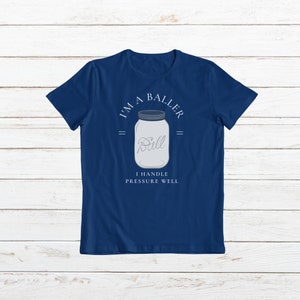 Puede incluir: Una camiseta azul marino con un gr&aacute;fico blanco de un frasco de vidrio con la palabra "Ball" en su interior. El texto "I'm a Baller" est&aacute; por encima del frasco y "I handle pressure well" est&aacute; por debajo del frasco.