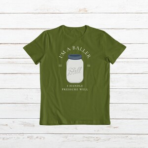 Puede incluir: Una camiseta verde con el texto "I'm a Baller" y un frasco de vidrio con la palabra "Ball" en su interior. El texto debajo del frasco dice "I handle pressure well."