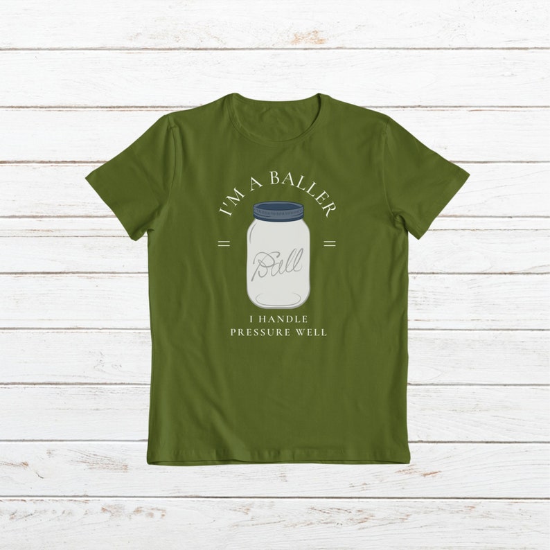 Puede incluir: Una camiseta verde con el texto "I'm a Baller" y un frasco de vidrio con la palabra "Ball" en su interior. El texto debajo del frasco dice "I handle pressure well."