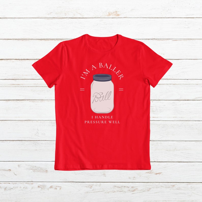 Puede incluir: Camiseta roja con un gr&aacute;fico blanco de un frasco de vidrio con la palabra "Ball" dentro. El texto alrededor del frasco dice "I'm a Baller" y "I handle pressure well".