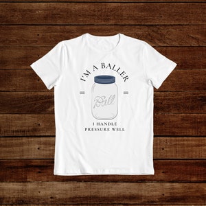 Puede incluir: Camiseta blanca con un gr&aacute;fico de un frasco de vidrio con la palabra "Ball" dentro. El texto "I'm a Baller" est&aacute; por encima del frasco y "I handle pressure well" est&aacute; por debajo del frasco.